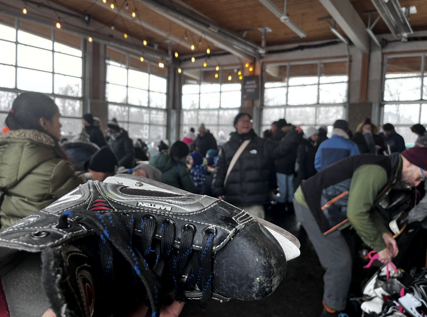 A hockey skate being held up in front of a packed room of people with twinkly lights and lots of windows.